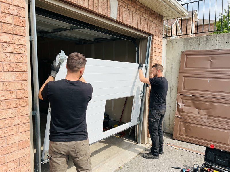 Two installers carrying a white door section toward a brick garage opening